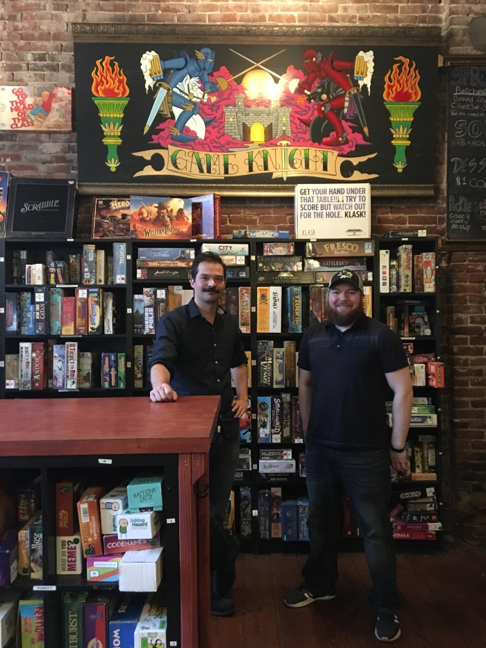 Two people in black shirts and pants stand in front of a wall of shelves containing board games.